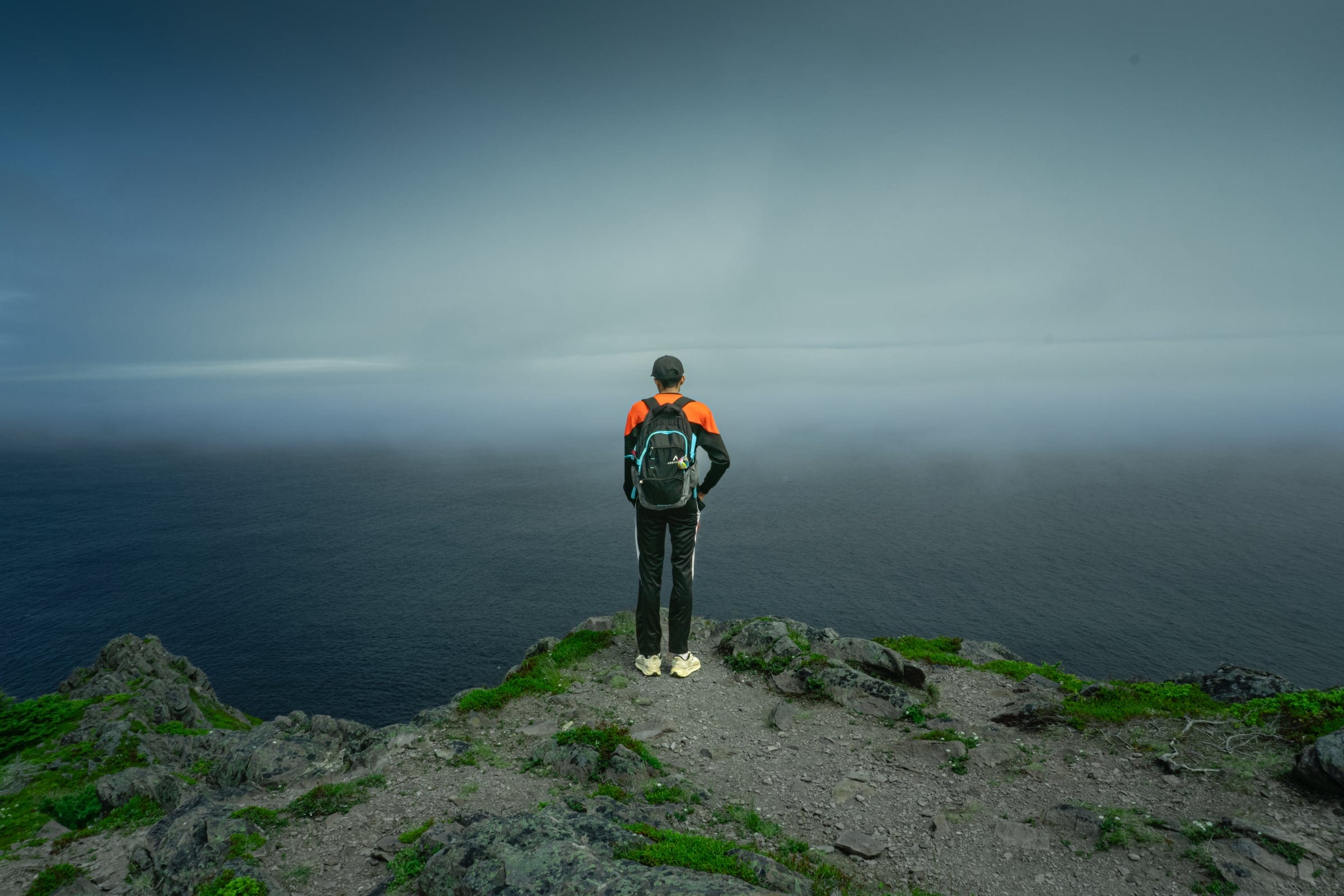 A hiker in an orange and black jacket stands on a rocky summit, facing a sea lost in grey fog.