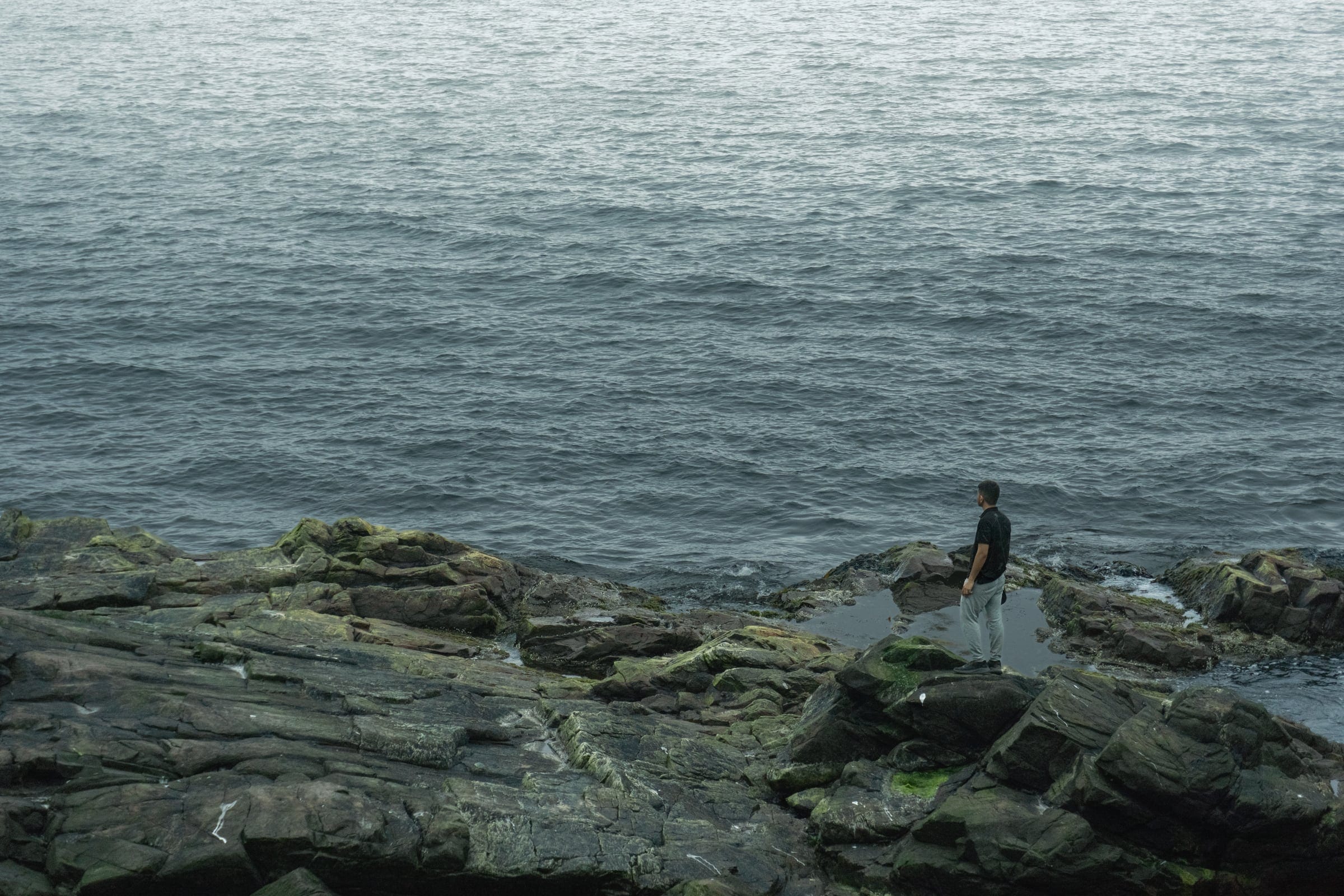 A lone figure on dark wet rocks at the edge of a grey, choppy sea.