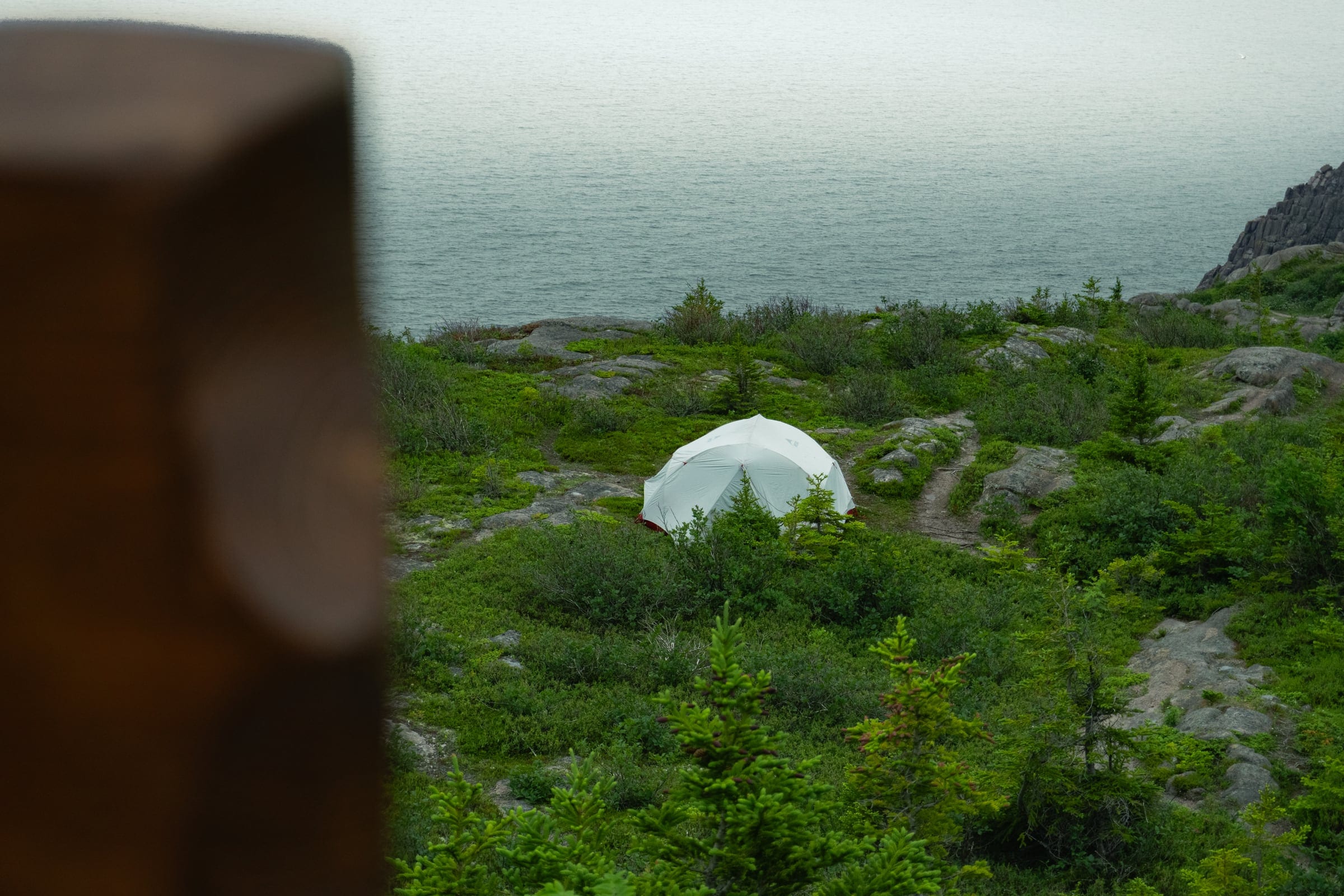 A small white dome tent in green scrub above the sea, a blurred wooden post in the foreground.