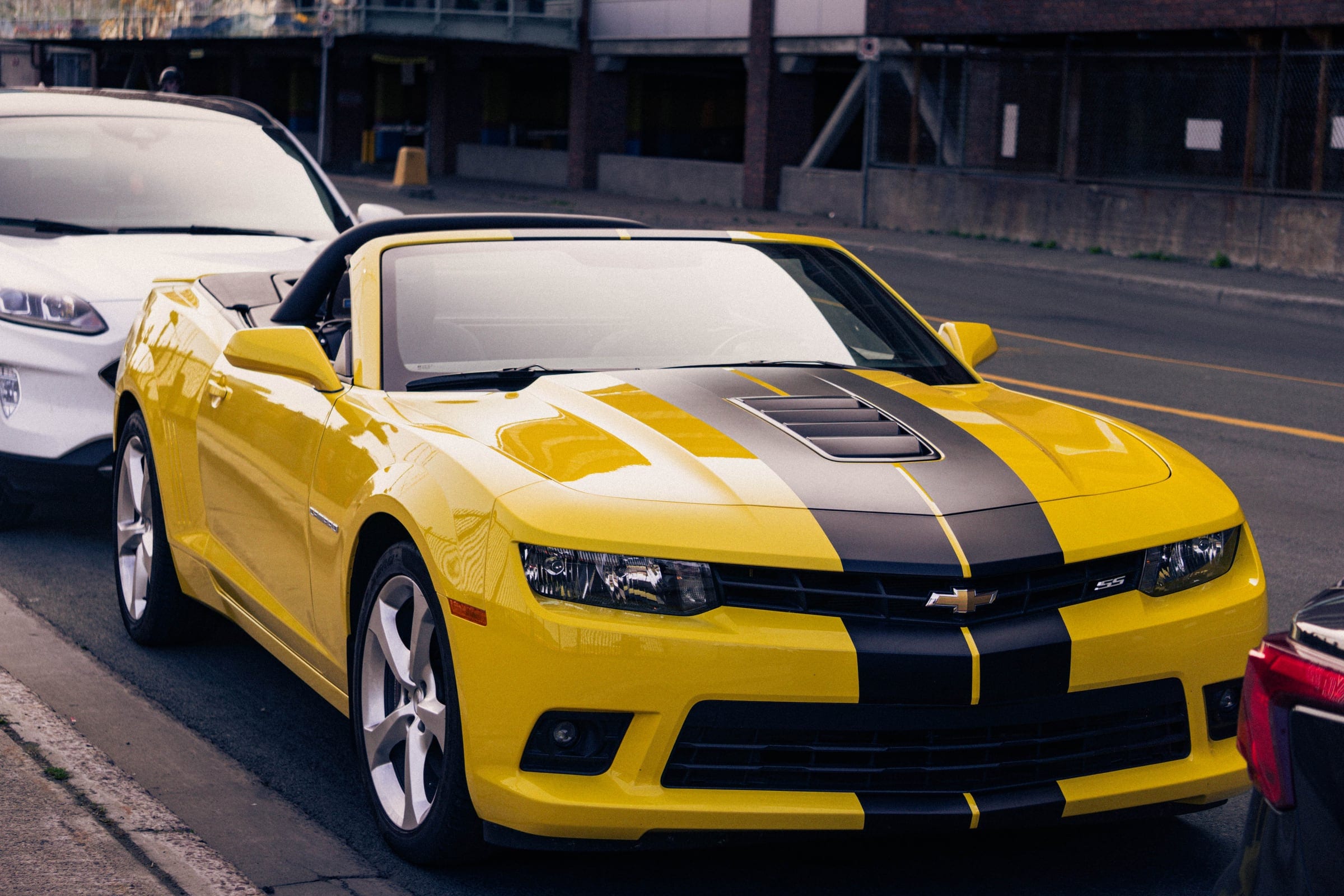 A yellow Chevrolet Camaro convertible with black racing stripes, parked at the kerb.