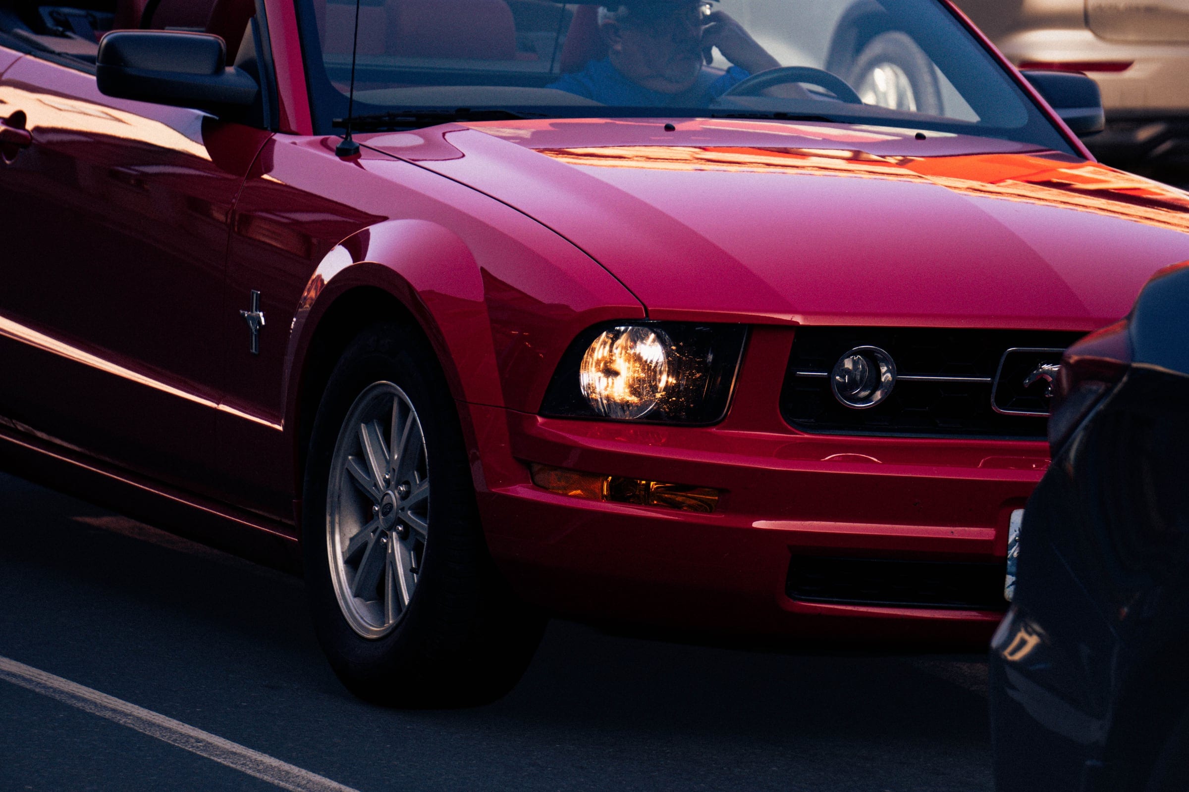 A red Ford Mustang convertible turning in low sunlight, headlight lit.