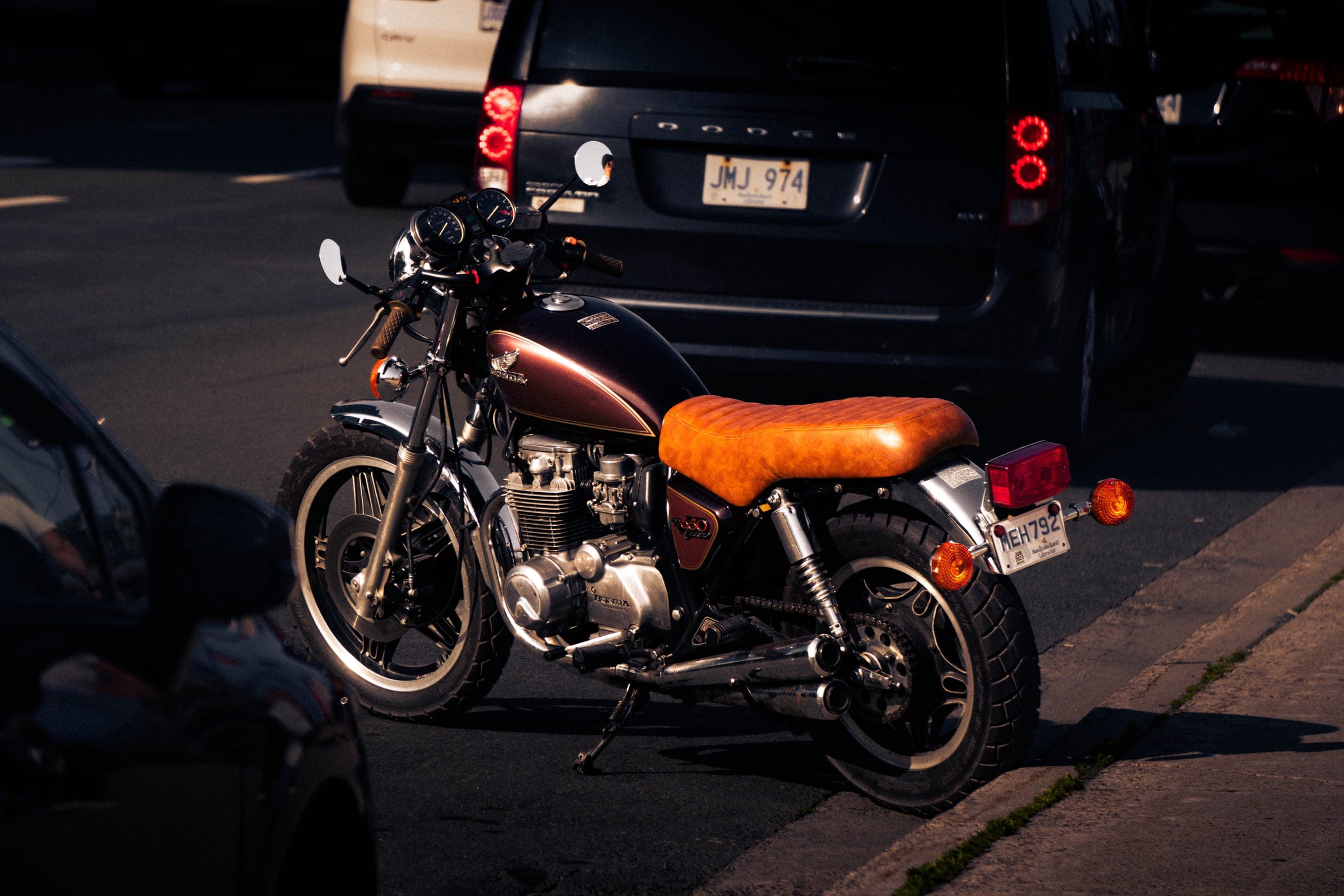 A vintage motorcycle with a tan leather seat parked at the kerb in evening shade.