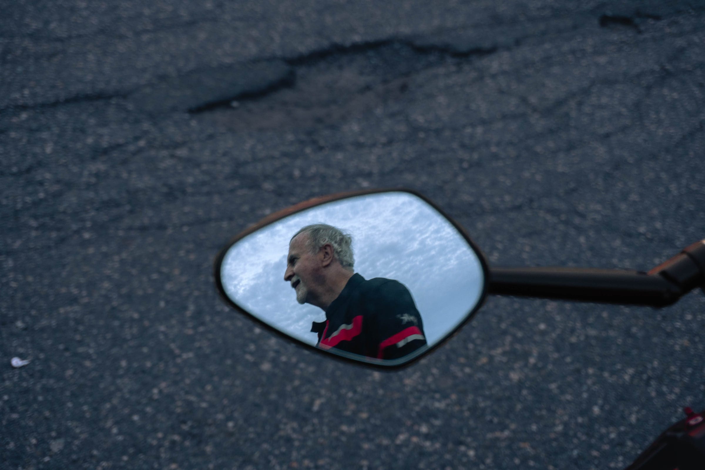 A rider’s face reflected in a motorcycle mirror, the road behind him.