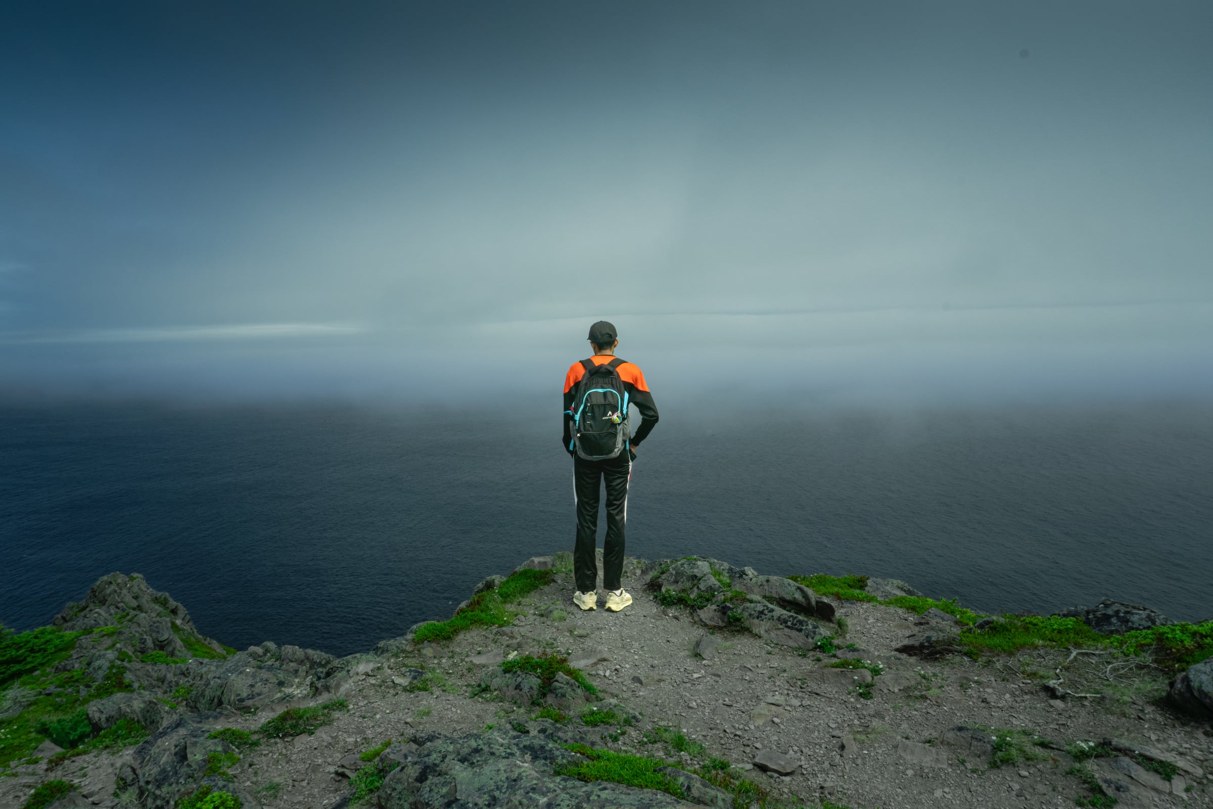 A hiker in an orange and black jacket stands on a rocky summit, facing a sea lost in grey fog.