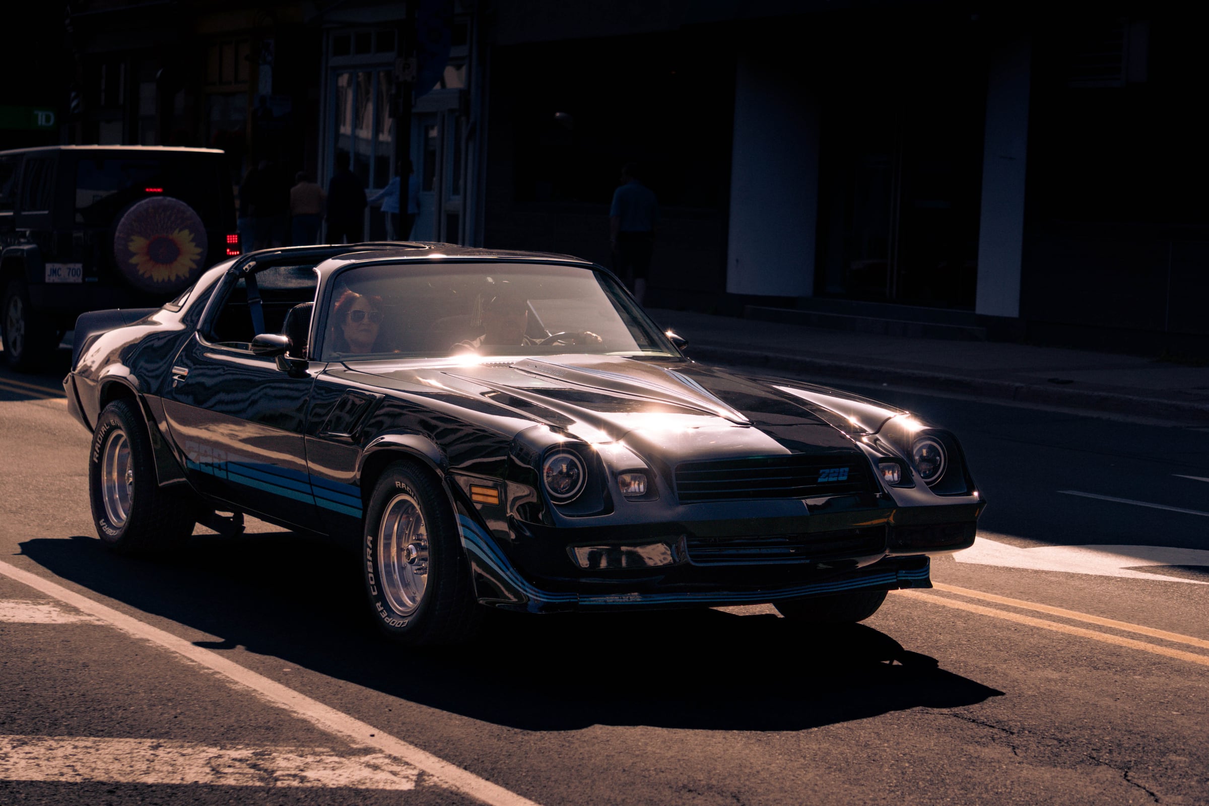 A black late-1970s Chevrolet Camaro Z28 driving through deep shade on a city street.