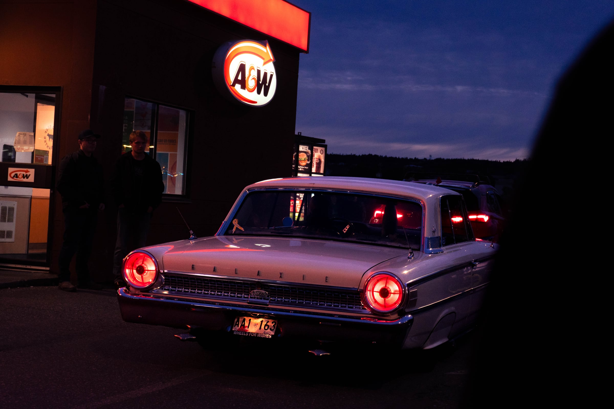 A classic car under an A&W sign after dark, its round tail lights burning red.